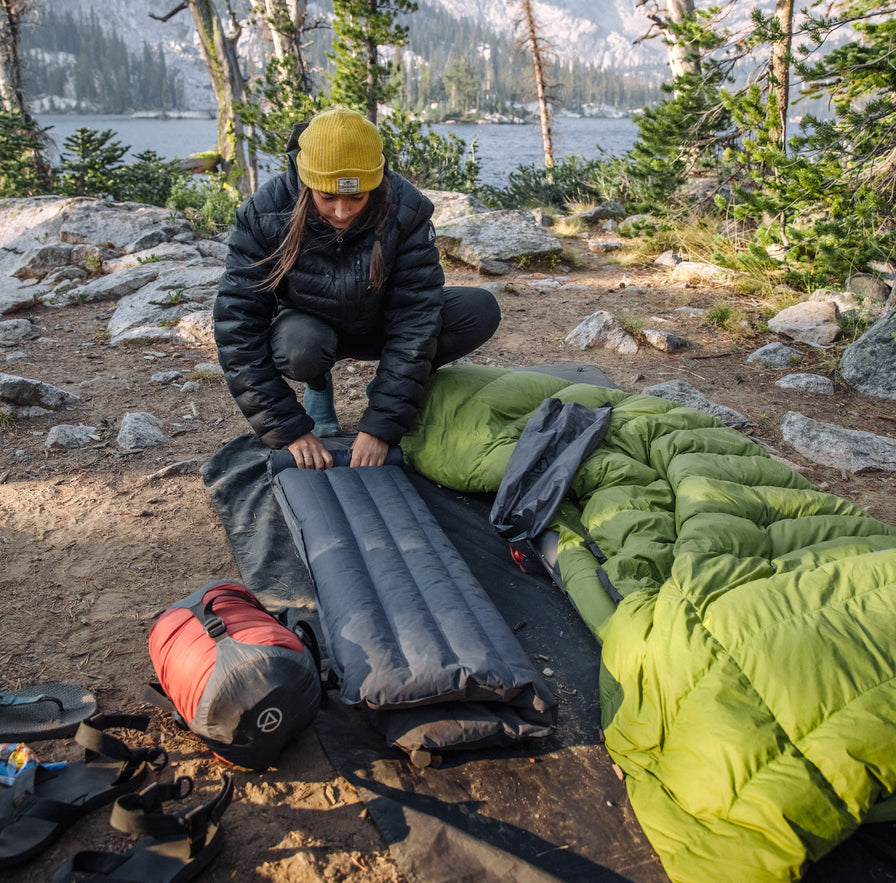 Camper setting up Zenbivy sleeping pad and green quilt at lakeside forest campsite, showcasing lightweight modular backpacking sleep system.